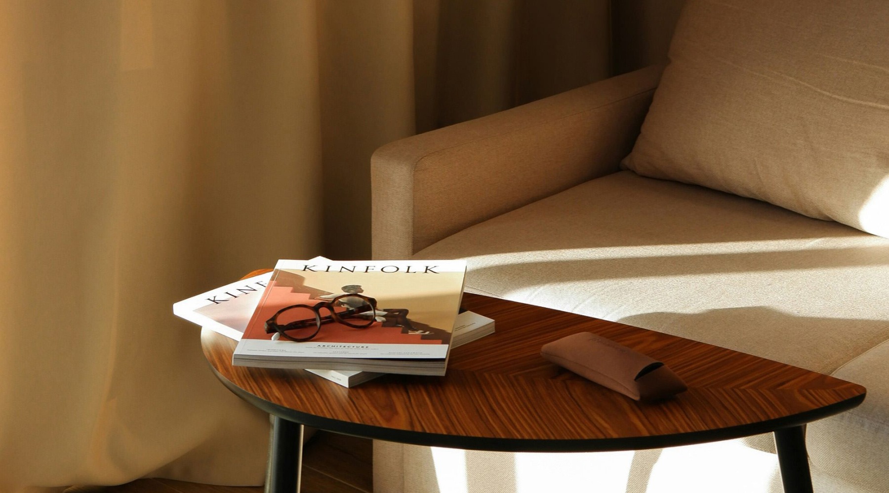 A table with a book and glasses on it next to a couch in the sunlight.